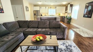 A modern living room with a sectional sofa, a coffee table with fruit, and a kitchen in the background.