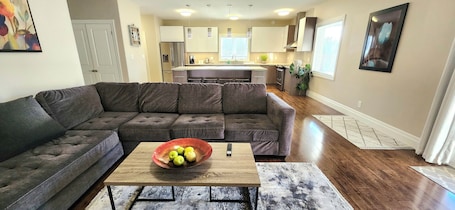 A modern living room with a sectional sofa, a coffee table with a bowl of fruit, and a kitchen in the background.. Spacious Doon Retreat | Sleeps 8 | Private Yard | Near 401