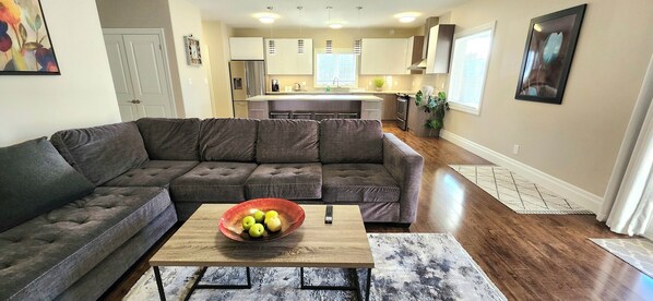 A modern living room with a sectional sofa, a coffee table with a bowl of fruit, and a kitchen in the background.