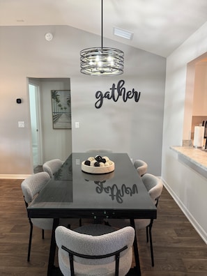 A modern dining room with a black table, grey chairs, and a 'gather' wall art piece.