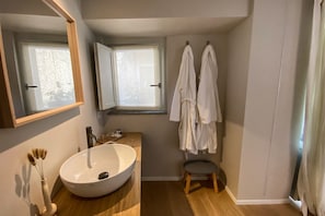 A bathroom with a large white sink, a mirror, a window, and two white robes hanging on the wall.