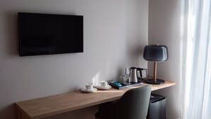Ein Hotelzimmer mit einem flachbildfernseher an der Wand, einem Holztisch mit schwarzem Stuhl, einer schwarzen Mülleimer und einer Tischlampe.