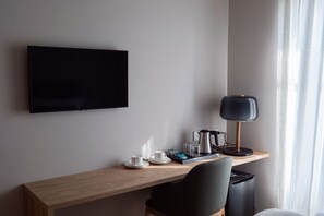 A hotel room with a flat-screen TV mounted on the wall, a wooden desk with a black chair, a black trash bin, and a table lamp.
