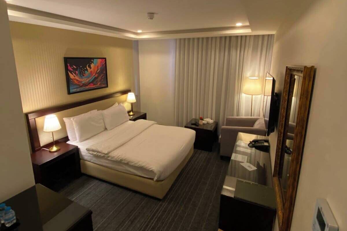 A hotel room with a bed, bedside tables, a desk, a chair, a mirror, a painting, and a window with curtains.