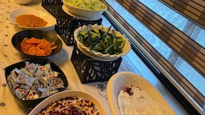 Una mesa de bufé con diversos platos, entre ellos ensaladas, verduras y postres, en una sala con un ventanal y persianas.
