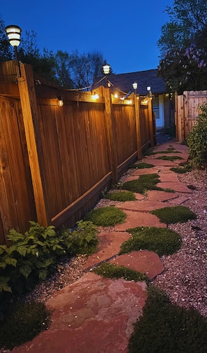 Ein Holzzaun mit Lichterketten, ein Kiesweg und viel Grün.