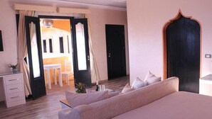 Quarto de hotel com cama, escrivaninha, cadeira e vista para um edifício através de uma porta aberta.