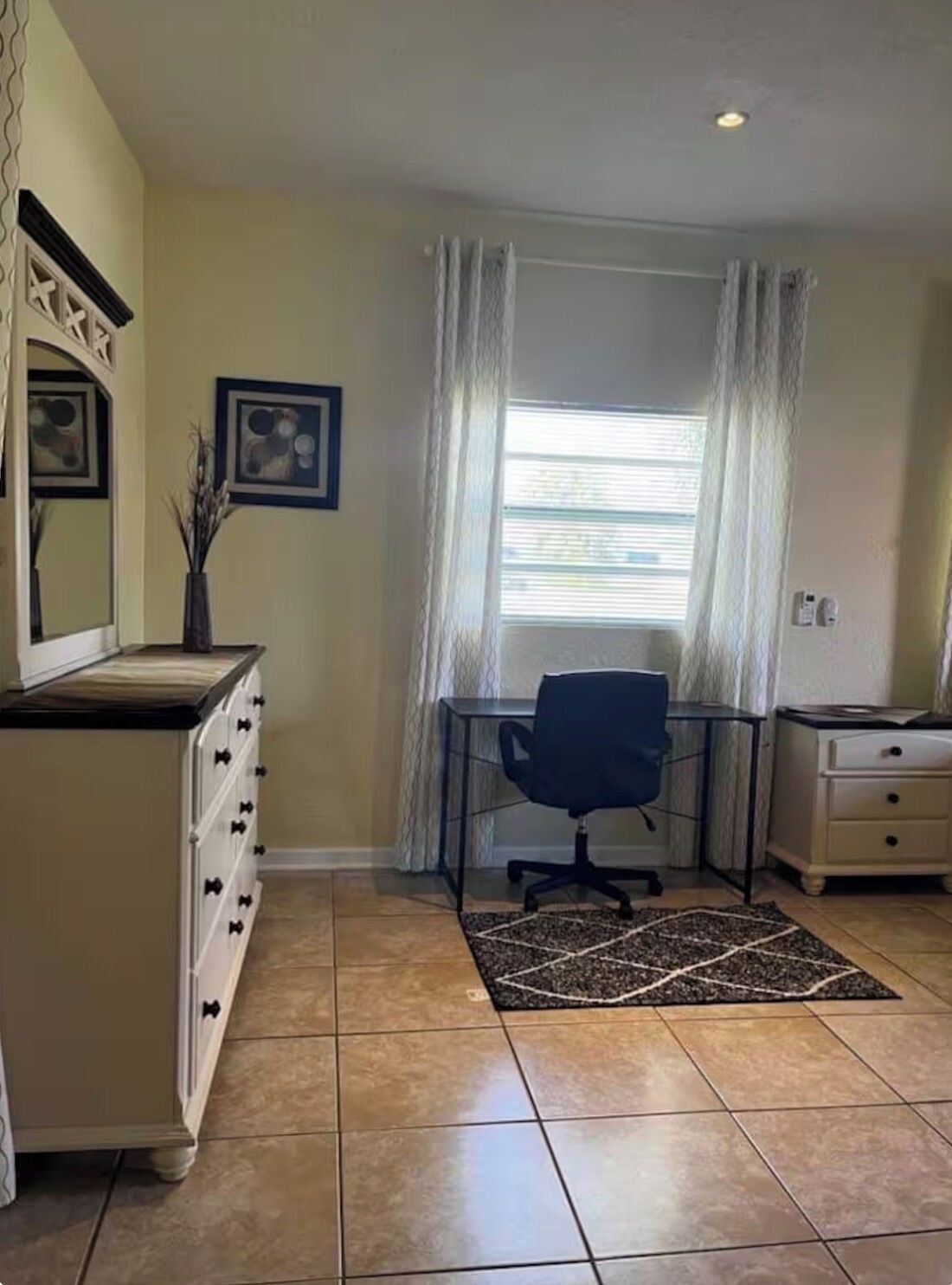 A room with a desk, chair, dresser, and a window with curtains.