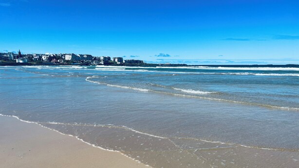 Ein Strand mit Wellen, blauem Himmel und Gebäuden im Hintergrund.