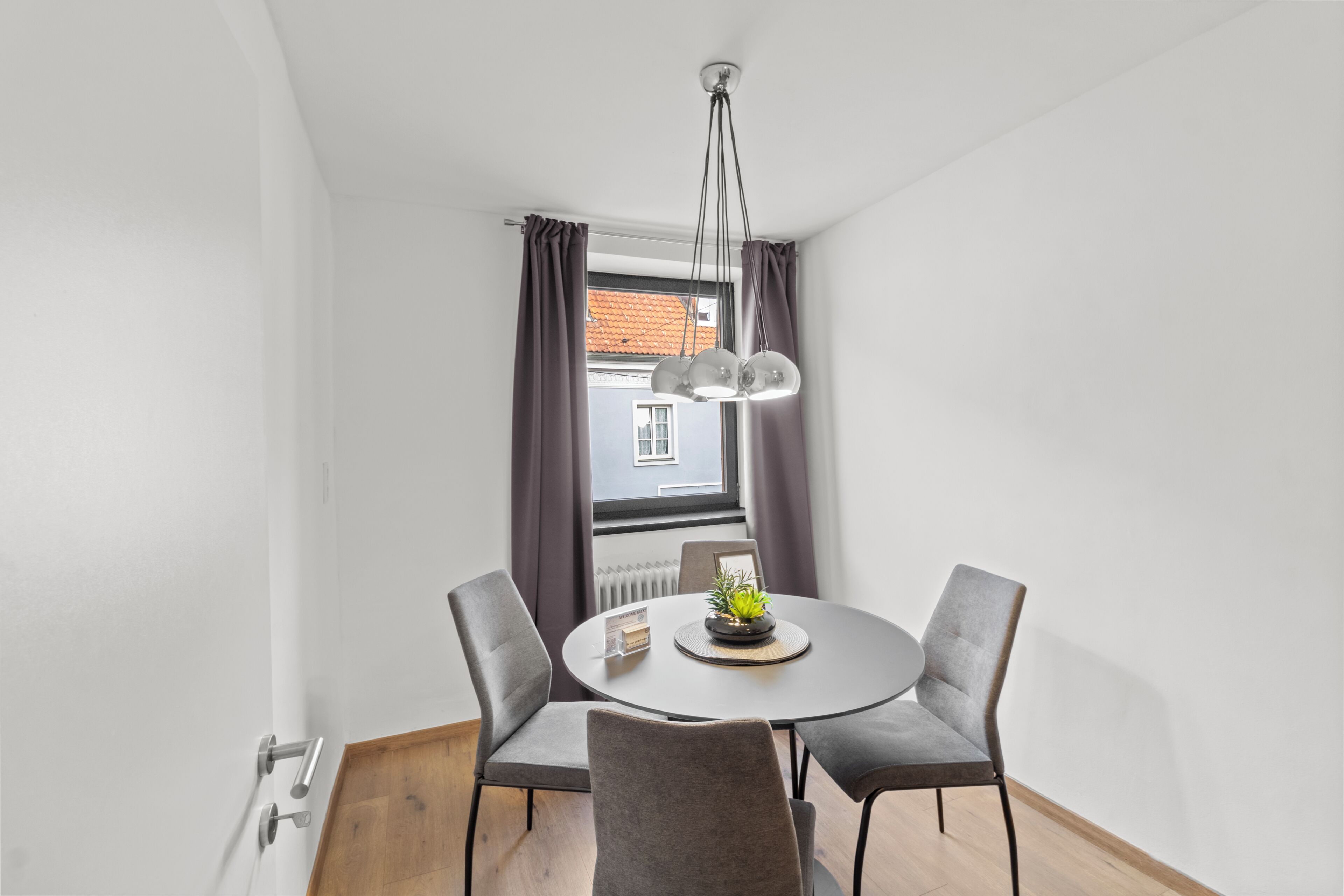 A modern dining area with a round table, four chairs, a pendant light, and a view of a building through a window with curtains.