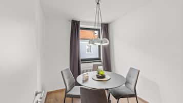 A modern dining area with a round table, four chairs, a pendant light, and a view of a building through a window with curtains.