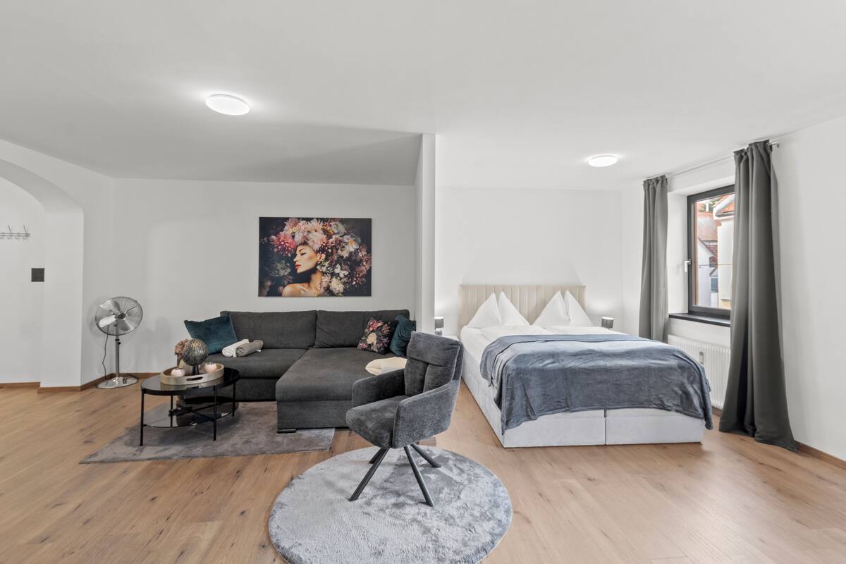 A modern room with a grey sofa, a round grey chair, a bed with grey bedding, a fan, and a painting on the wall.