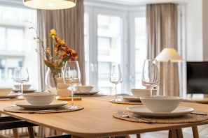 Ein Esstisch ist gedeckt mit weißen Schüsseln und Weingläsern, eine Vase mit Blumen und eine Lampe in einem Raum mit großen Fenstern und Vorhängen.