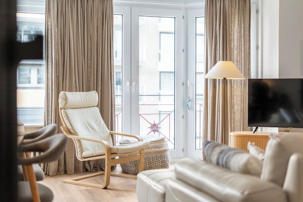 Ein Wohnzimmer mit einer Couch, einem Schaukelstuhl, einem Fernseher und einem Balkon mit Vorhängen.