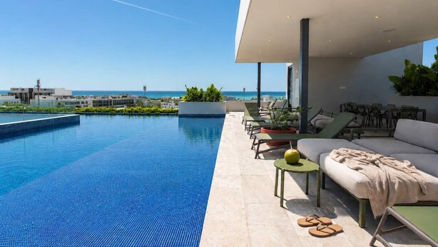 A modern outdoor pool area with lounge chairs and a sofa, overlooking a clear blue sky and a distant view of buildings. - Ceiba 25 | Rooftop Pool, Gym & 5th Ave Condo (Playa del Carmen)