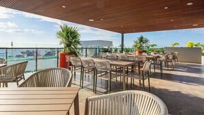 An outdoor patio with tables and chairs, potted plants, and a view of the ocean. - Ceiba 25 | Rooftop Pool, Gym & 5th Ave Condo (Playa del Carmen)