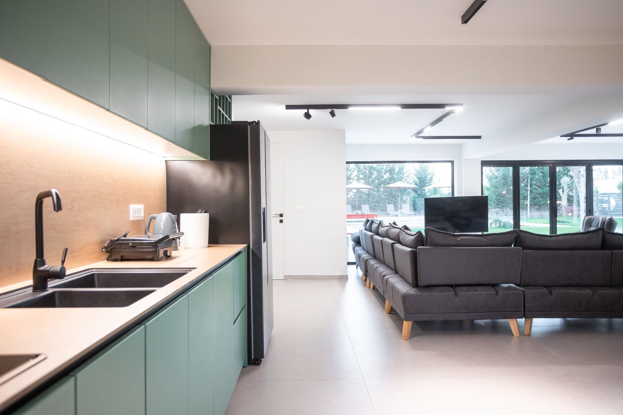 A modern kitchen with a built-in stove, a sink, and a countertop. There is a dark-colored sofa in the living area with a flat-screen TV.