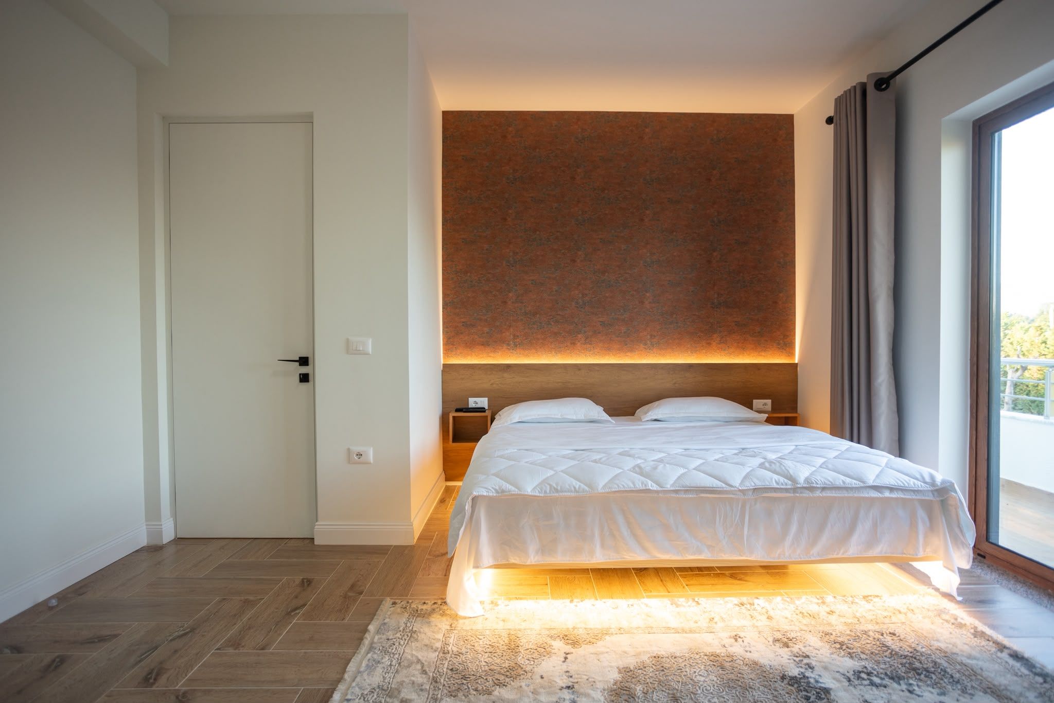 Une chambre moderne avec un grand lit, une tête de lit en bois, une table de chevet et une vue sur l’extérieur par une fenêtre.