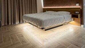 Une chambre d’hôtel moderne avec un parquet en bois disposé en chevrons, un grand lit avec une tête de lit, un luminaire au plafond et une fenêtre avec des rideaux.