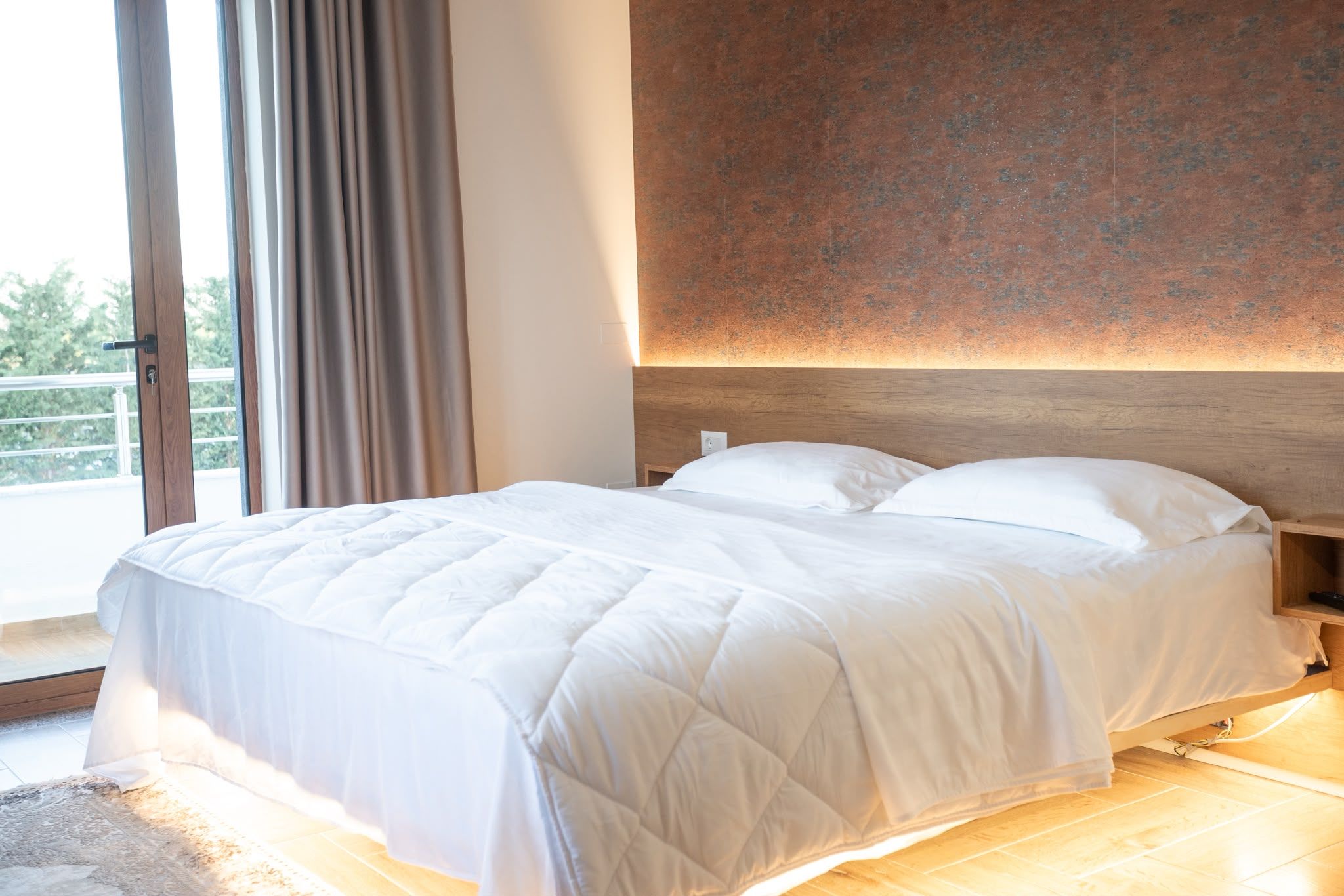 A hotel room with a large bed, a wooden headboard, and a view through a window with curtains.