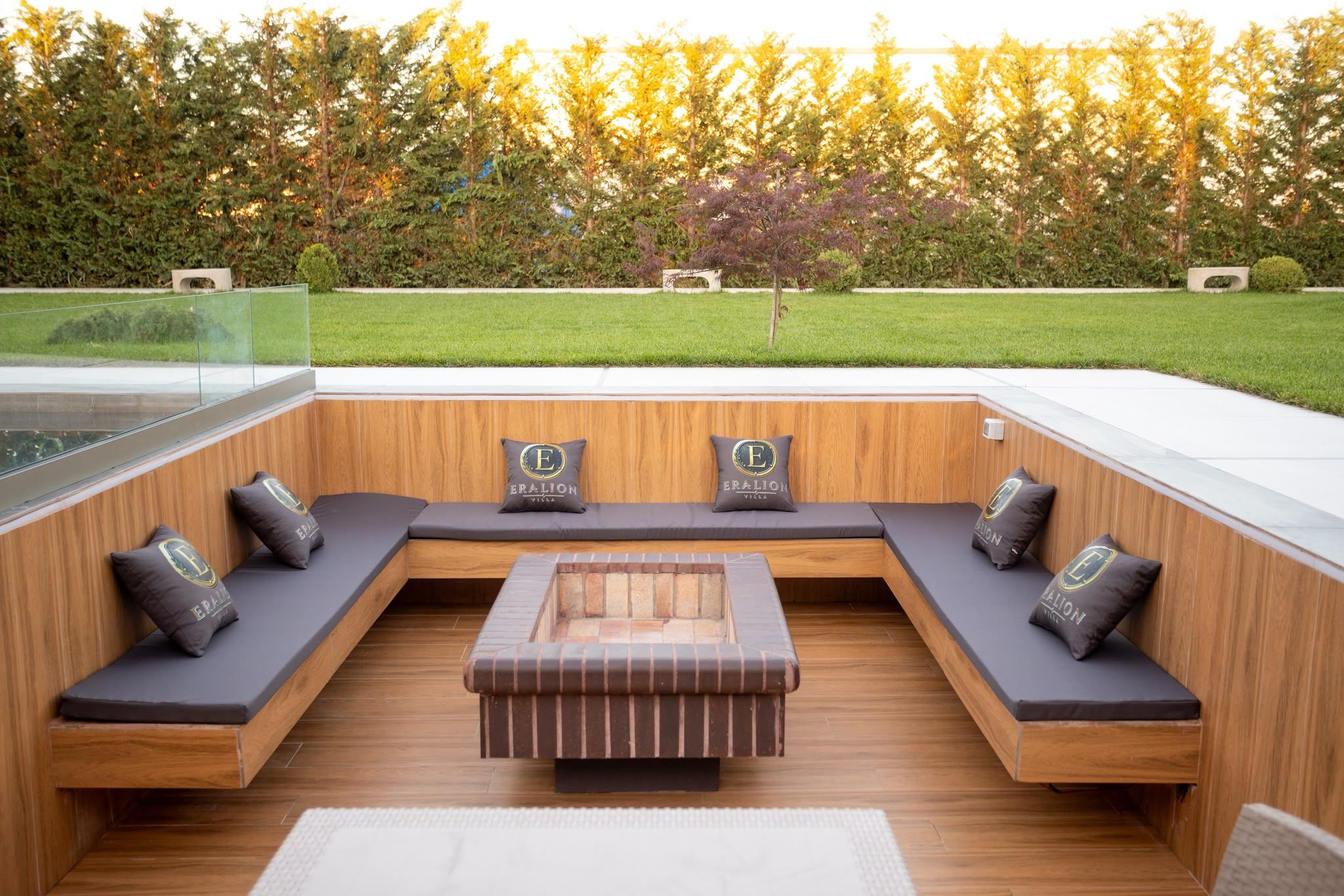 A modern outdoor seating area with a wooden bench and cushions, a central table, and a view of a well-maintained garden and trees.