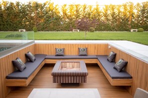 Un espace extérieur moderne, aménagé pour se détendre, avec un banc en bois et des coussins, une table centrale, et une vue sur un jardin et des arbres bien entretenus.