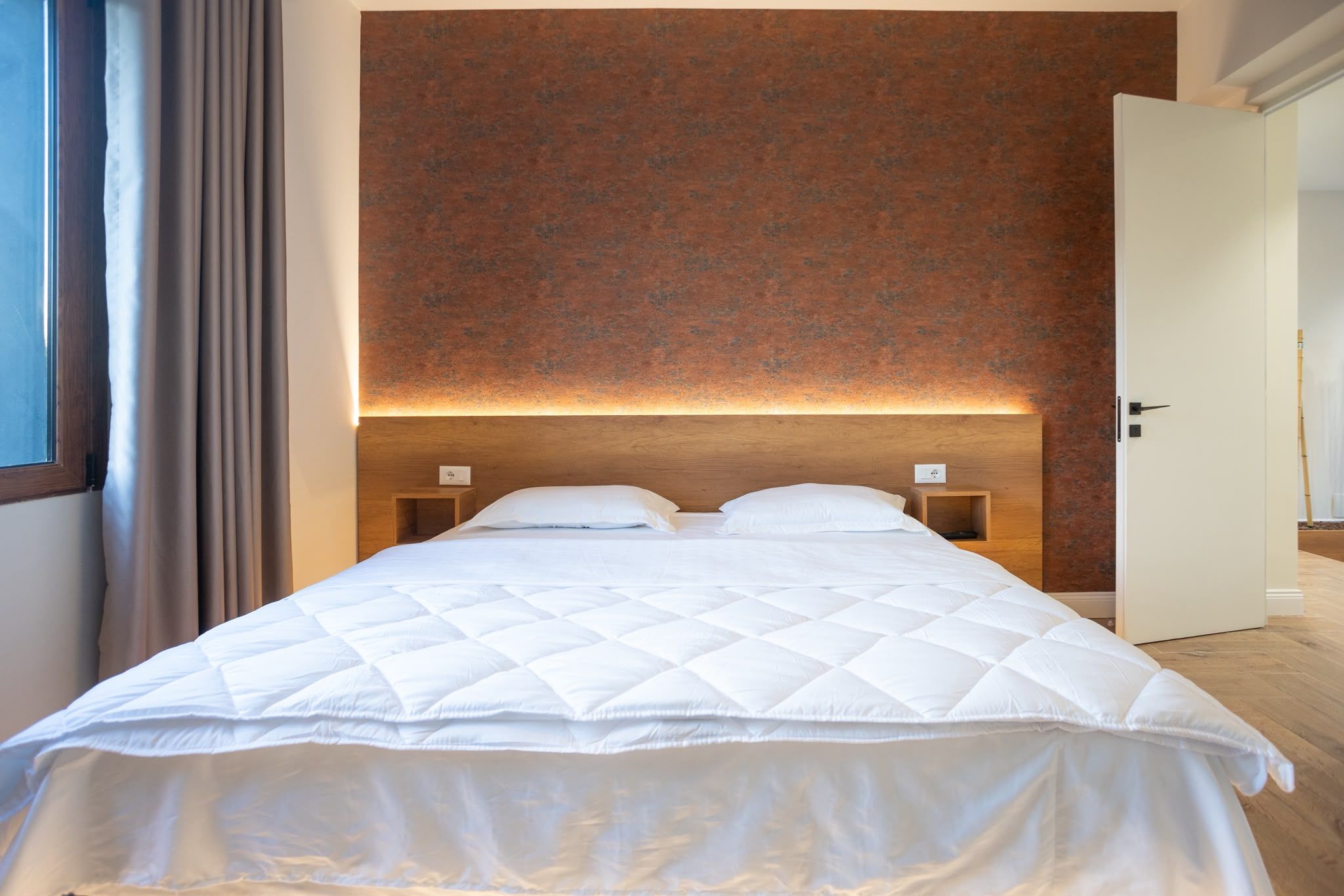 Cama bem arrumada com edredom branco acolchoado em quarto com parede marrom texturizada e cabeceira de madeira.