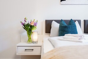 Een netjes opgemaakt bed met kussens, een nachtkastje met een bloemenvaas en een lamp.
