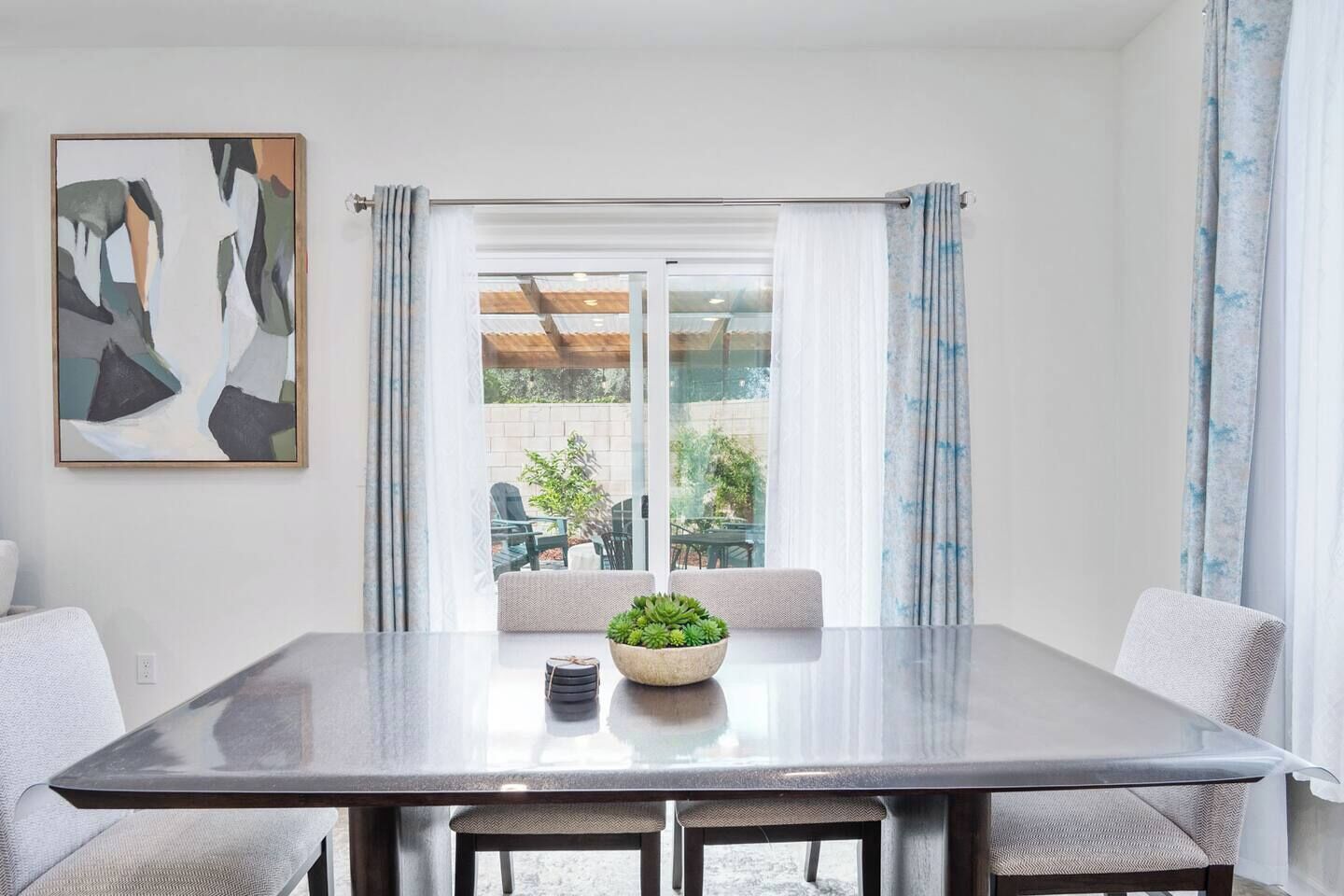 Un espace repas moderne avec une table en verre, des chaises blanches, une plante en pot et, à travers une porte vitrée, une vue sur un coin salon extérieur.