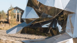 Una escultura metálica geométrica con superficie reflectante, ubicada en un paisaje natural con un edificio al fondo.
