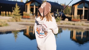 Una persona pescando junto a un cuerpo de agua tranquilo con cabañas de madera al fondo.