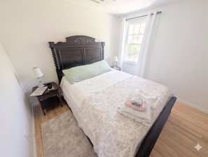 Een keurig opgemaakt bed met hoofdbord, nachtkastje met lamp en een raam met gordijnen.