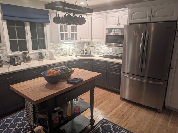 Een keuken met een roestvrijstalen koelkast, een houten tafel met een fruitschaal en een hangende pannendroger.