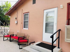 A house with a porch, two wicker chairs with red cushions, a white door, and a small white fence.