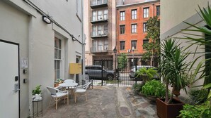 A narrow courtyard with stone flooring, a small seating area with chairs and a table, potted plants, and a building with multiple windows and balconies in the background.