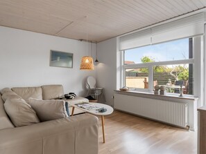 Ein modernes Wohnzimmer mit einer Couch, einem Tisch, einem Sessel, einer Pendelleuchte und einem Fenster mit Blick ins Freie.