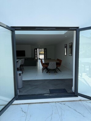 Une porte-fenêtre coulissante donne accès à un intérieur moderne comprenant un coin repas, un canapé et un téléviseur.