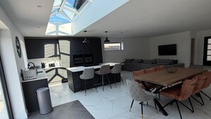 Cozinha moderna com clarabóia, área de jantar com mesa e cadeiras grandes e sala de estar com sofá e TV.