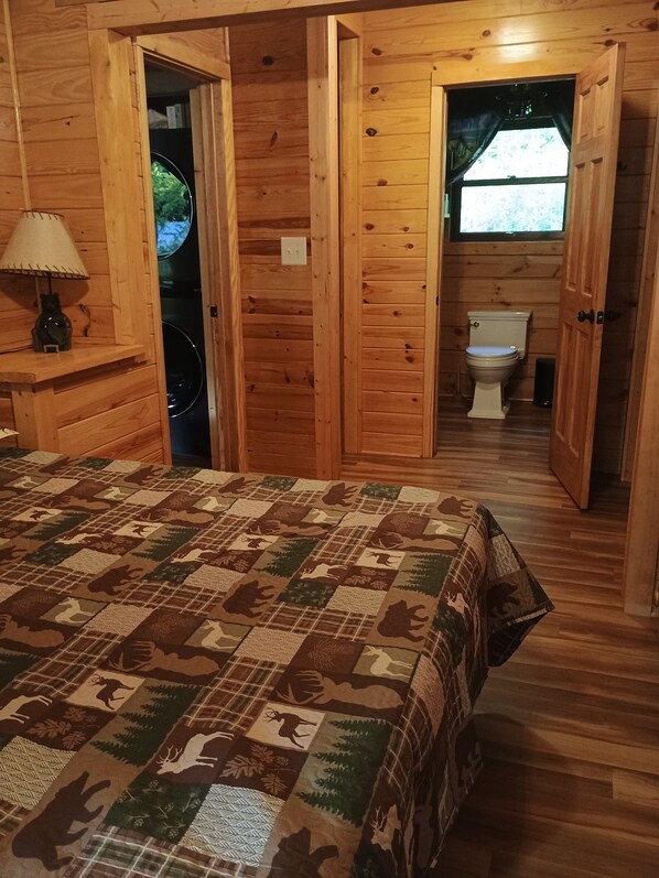 A wooden cabin bedroom with a quilted bedspread, a lamp, and a view of the outdoors through a window. - Cozy, secluded Dunlap cabin with a creek and playset by hangglider landing zone (Dunlap)
