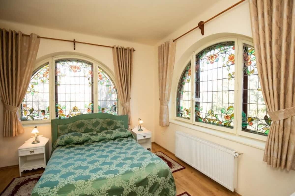 A bedroom with a bed, two bedside tables, a nightstand, a radiator, and two large arched windows with stained glass.