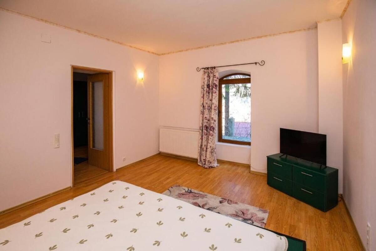 A bedroom with a bed, a television on a dresser, a window with curtains, and a door leading to another room.