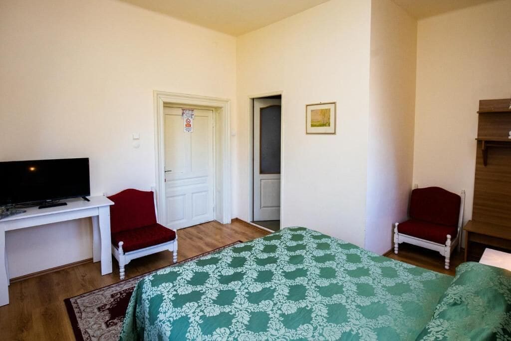A bedroom with a bed, two chairs, a desk with a television, and a door.