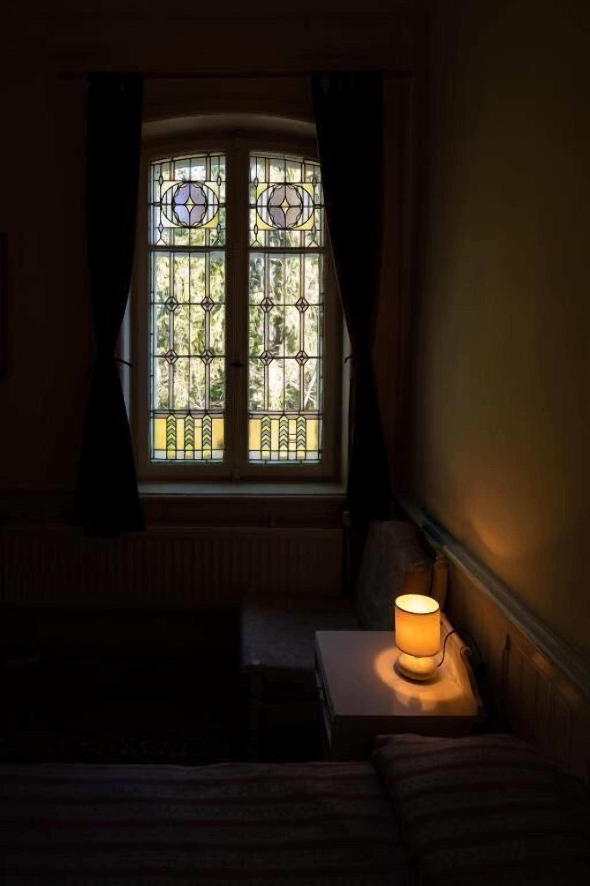 Una habitación con ventana, una lámpara en la mesita de noche y una cama.