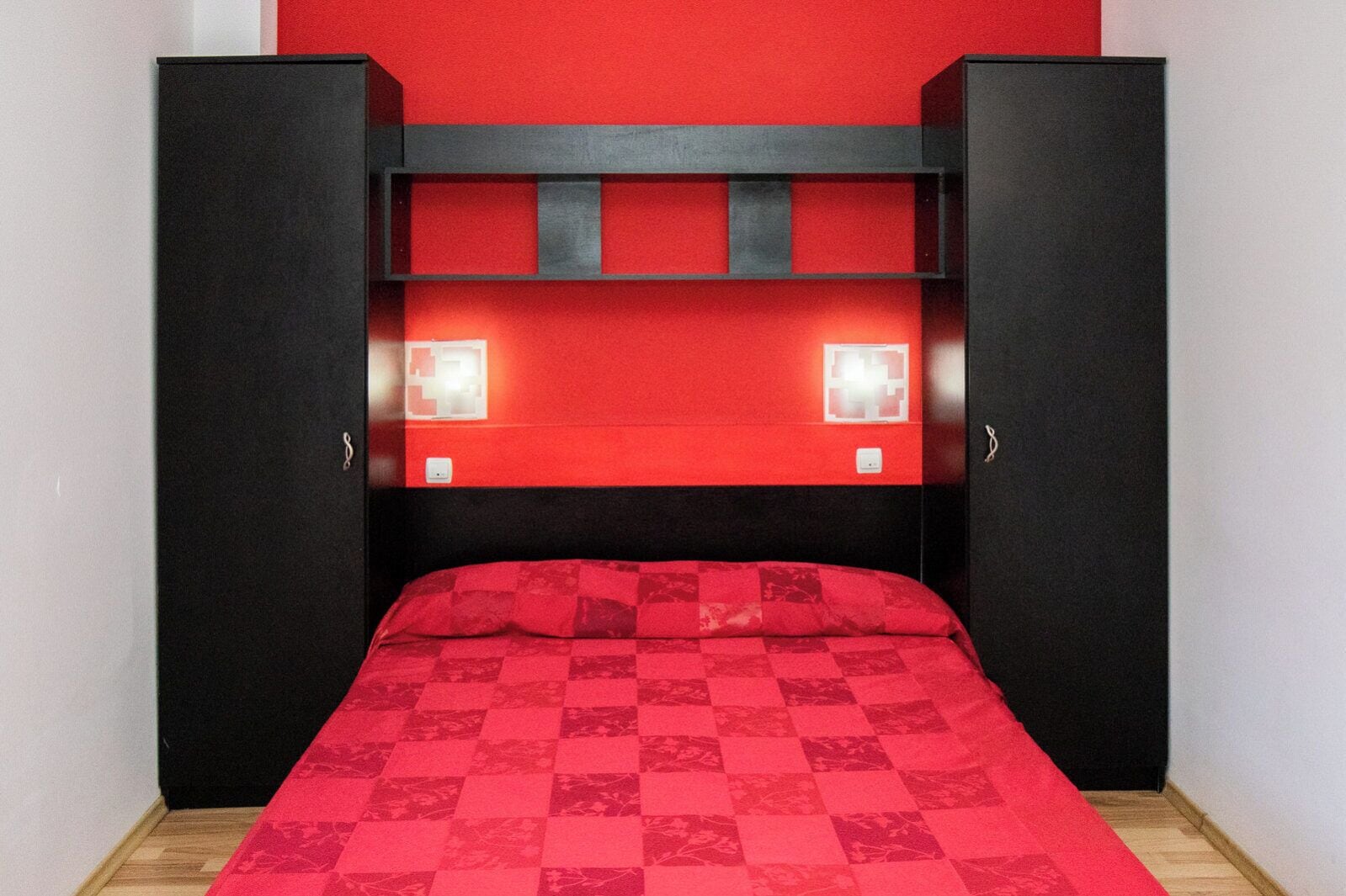 A room with a bed, black wardrobe, red wall, and two wall-mounted lights.