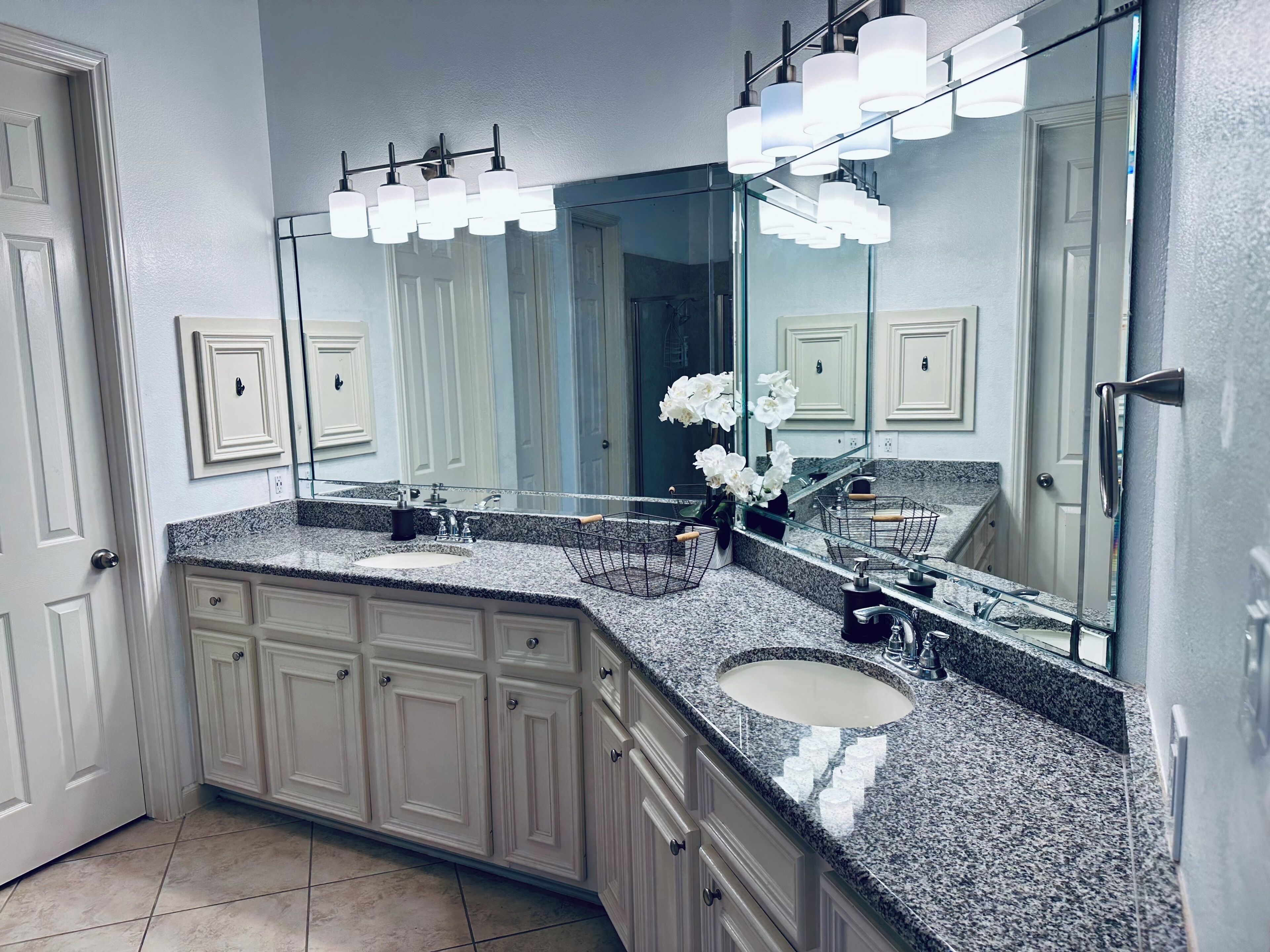 Un baño con encimera de granito, lavamanos doble, armarios blancos, un espejo grande y un jarrón con flores blancas.