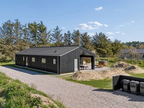 Un bâtiment moderne avec une allée en gravier, un espace extérieur aménagé et couvert, et un paysage boisé en arrière-plan.