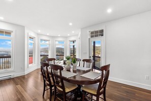 A dining room with a round table, six chairs, and a view of the outdoors through large windows.