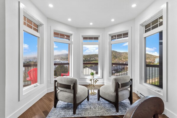 A corner room with large windows, two chairs, a small table, and a view of a hilly landscape.