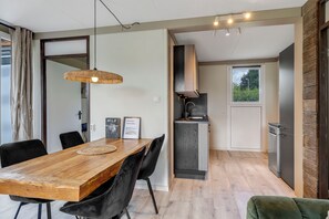 A modern kitchen with a built-in oven, a small window, and a dining area with a wooden table and black chairs. - Bungalow with large private garden and BBQ place near Lake Veere (Kortgene)