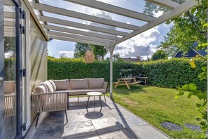 A covered patio with a sofa and table, a wooden bench, and a view of a hedge and house. - Bungalow with large private garden and BBQ place near Lake Veere (Kortgene)